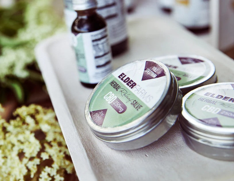 Salve containers with visible branding on a tray surrounded by elder flowers.