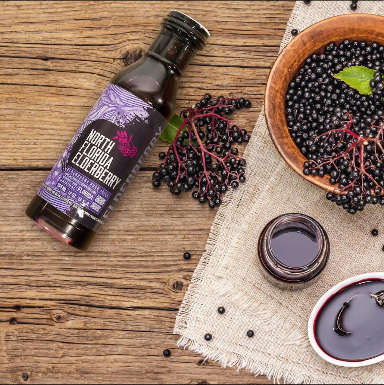 North Florida Elderberry Pure Juice on a wooden surface with elderberries and a bowl of berries.