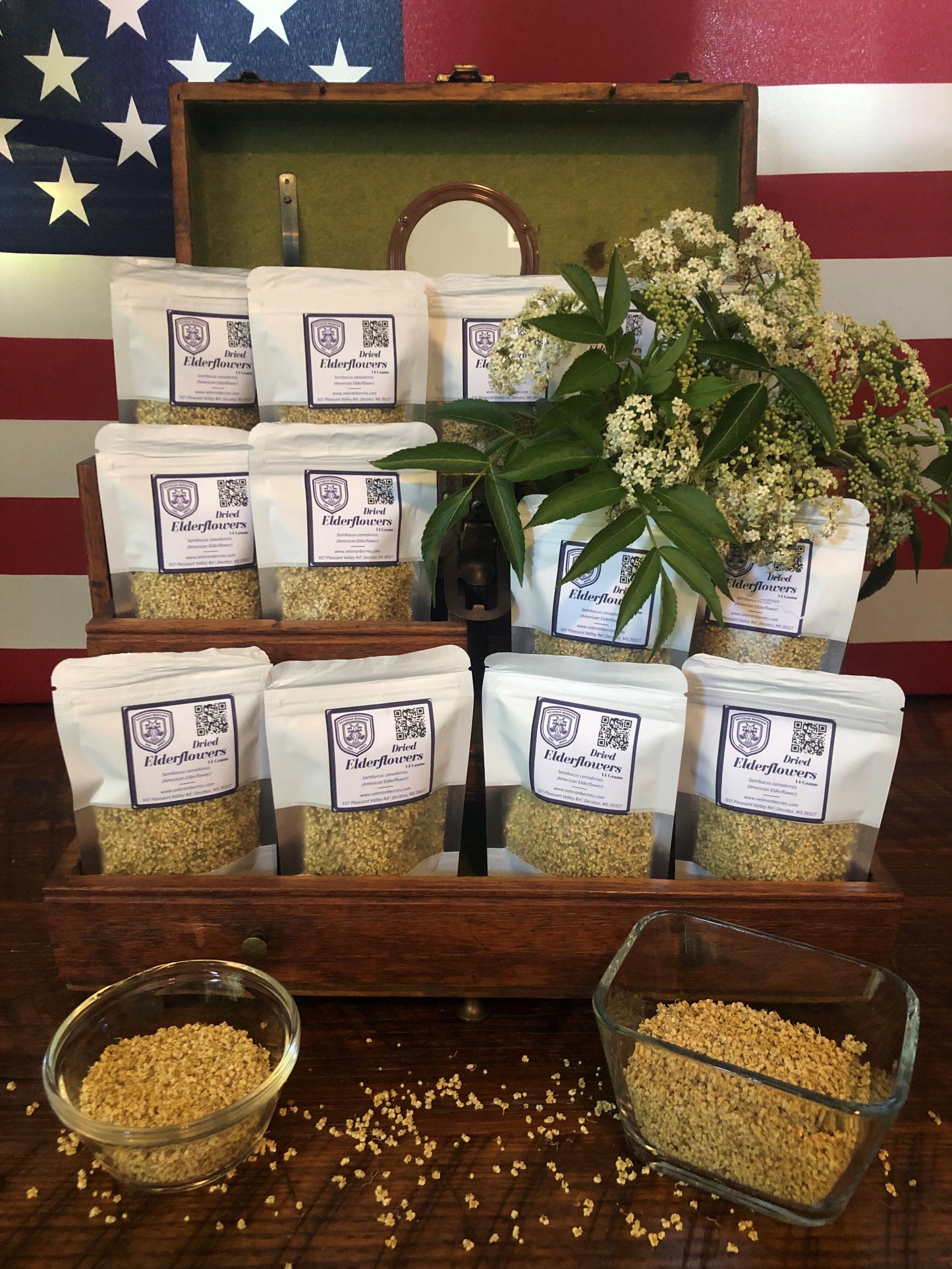 Veteran Berries Dried Elderflowers in wooden box on a wood table with dried elderflowers in two glass containers with sprinkles of elderflowers surrounding with the US Flag as backdrop.