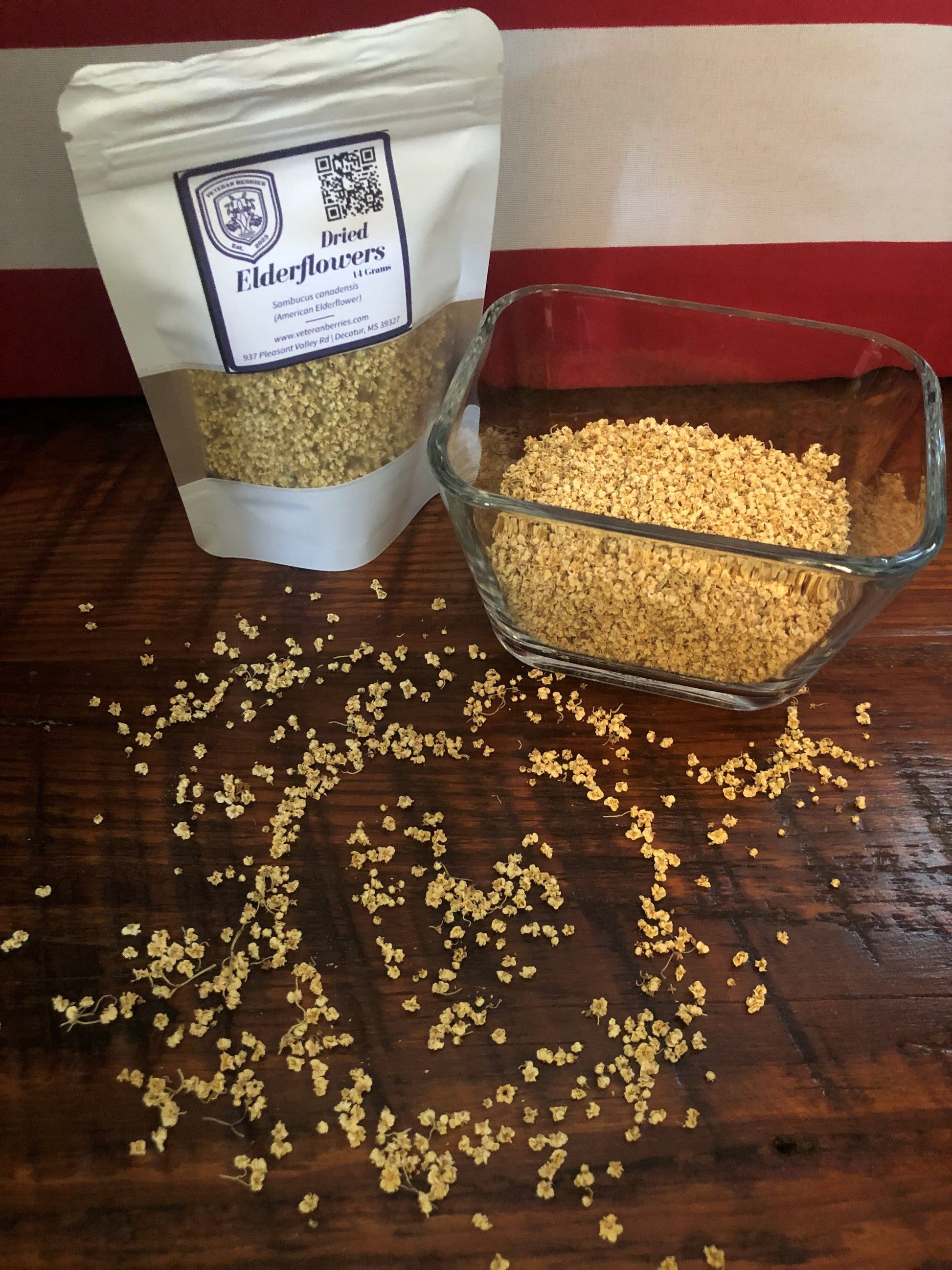 Dried elderflowers in a glass bowl and packaging on a wooden surface with spilled elderflowers on wood surface in shape of a heart and US Flag as backdrop.
